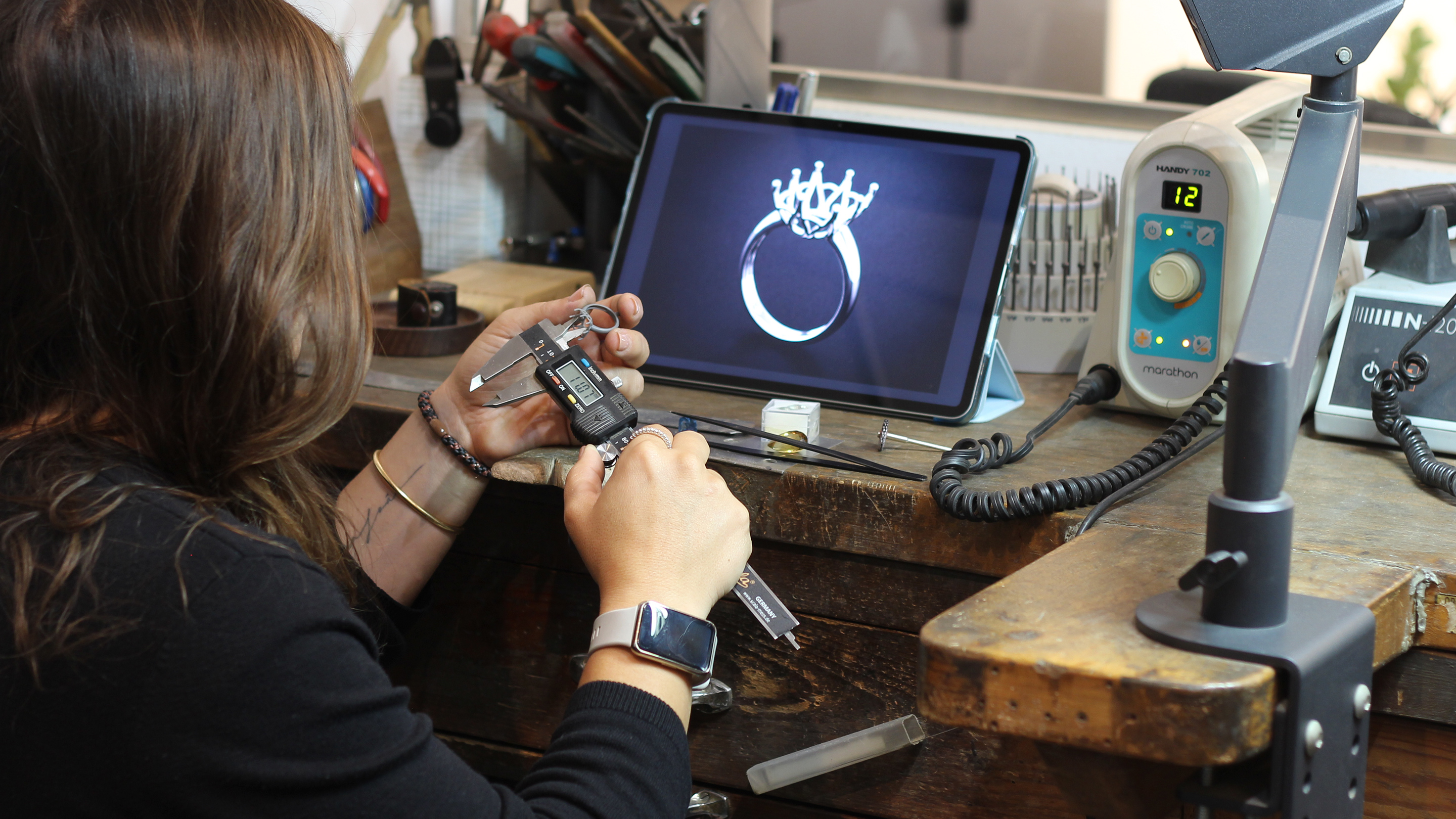 Diana at a wooden bench checking a prototype with callipers while a tablet shows the CAD render of the same piece